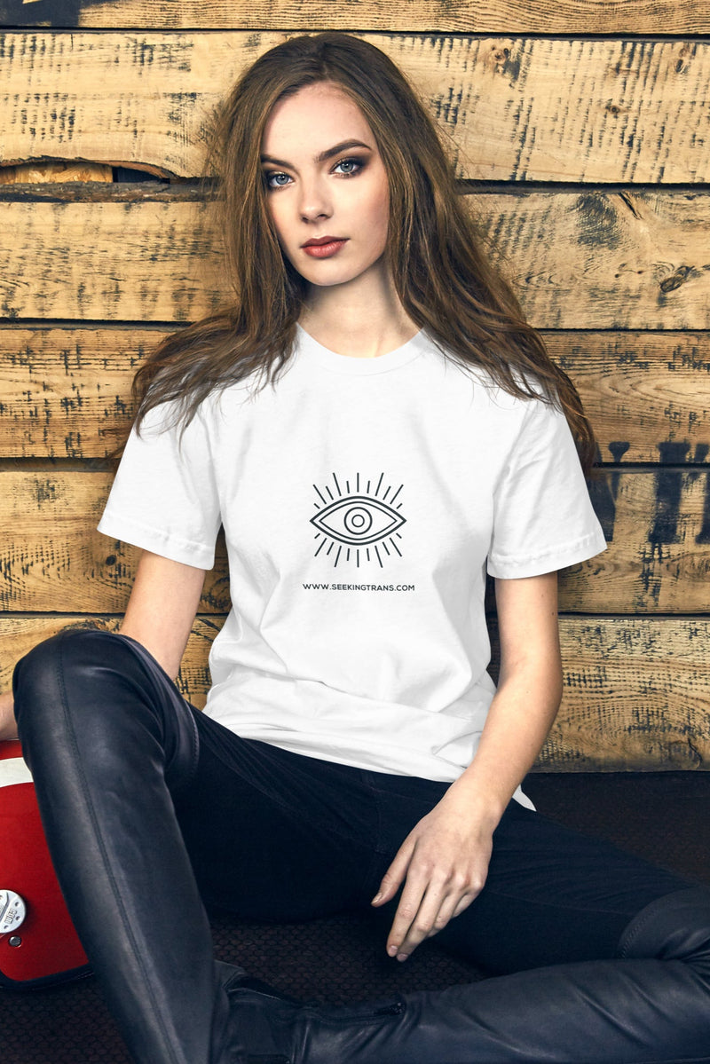 Model wearing white unisex t-shirt with Seeking Trans classic eye logo, sitting beside a red helmet and wearing black trousers
