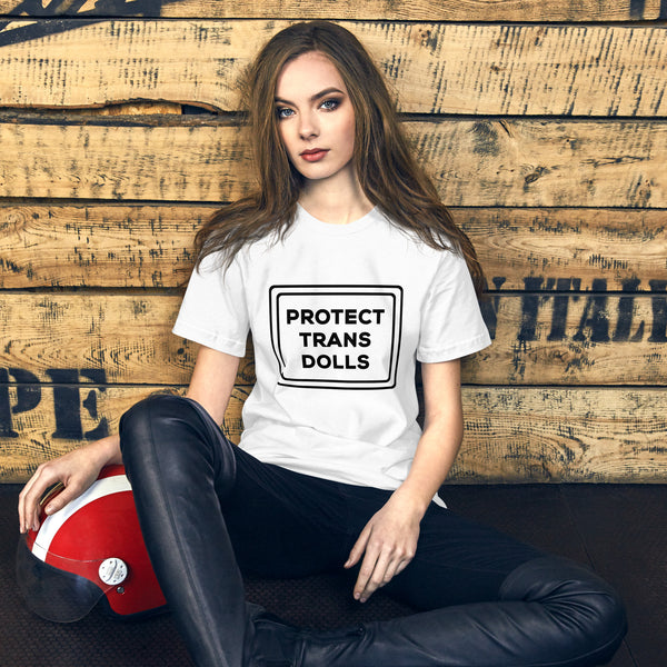 Model in white unisex Protect Trans Dolls t-shirt, sitting beside a red helmet and wearing black trousers