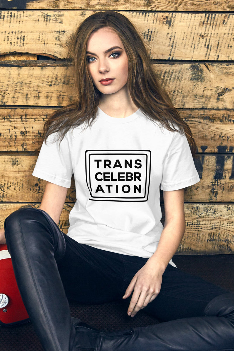 Person sitting casually beside a red helmet, wearing a white "Trans Celebration" T-shirt with bold black print, against a wooden backdrop